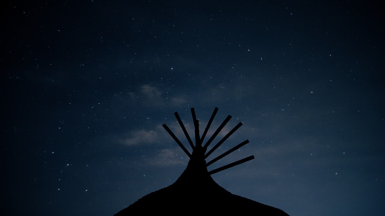 Early evening of a starry dark blue slightly cloudy sky with the silhouette of a structure's roof that comes to a point with eight spokes radiating from the top